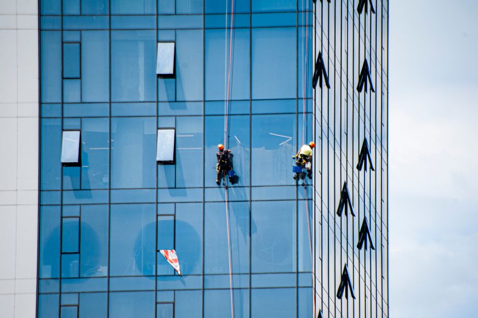 Two window cleaners working on a tall modern glass skyscraper on a clear day, showcasing skilled labor and urban architecture.