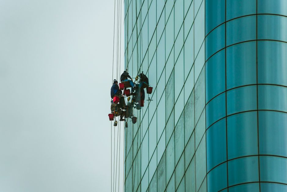 Window cleaners suspended on ropes working on a tall glass building facade.